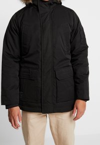 Černá zateplená parka s kapucí podšitým kožešinou, která má několik předních kapes, zapínání na zip a odolnou texturu látky.