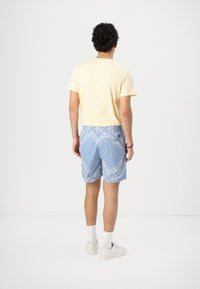Gul kortermet skjorte kombinert med blå mønstrete shorts. Shortsene har intrikate hvite design, elastisk midje og sidelommer. Hvite joggesko.