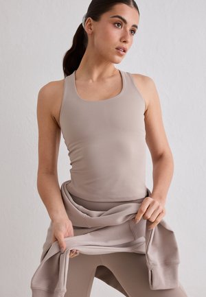 Femme aux cheveux foncés attachés en queue de cheval, portant un débardeur beige et un legging, tenant un sweat beige autour de la taille, regardant sur le côté.