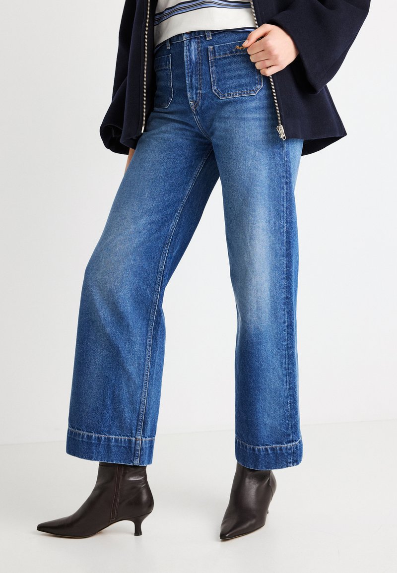Φαρδιά μπλε τζιν από τζιν ύφασμα, με ψηλή μέση, παραδοσιακό σχέδιο με πέντε τσέπες και ξεφτισμένο τελείωμα, σε συνδυασμό με σκούρα μποτάκια με τακούνι στο αστράγαλο.