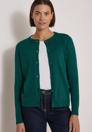 Femme portant un cardigan vert boutonné par-dessus une chemise blanche et un jean bleu foncé, debout devant un fond clair uni.