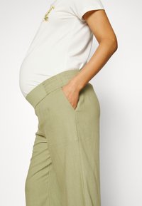 Calças de maternidade em linho verde claro com um cós elástico e bolsos laterais, combinadas com uma t-shirt branca de manga curta.