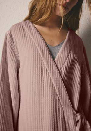 Femeie cu un halat roz prăfuit texturat peste un top gri, cu părul lăsat liber care îi acoperă parțial fața, în fața unui perete deschis la culoare.