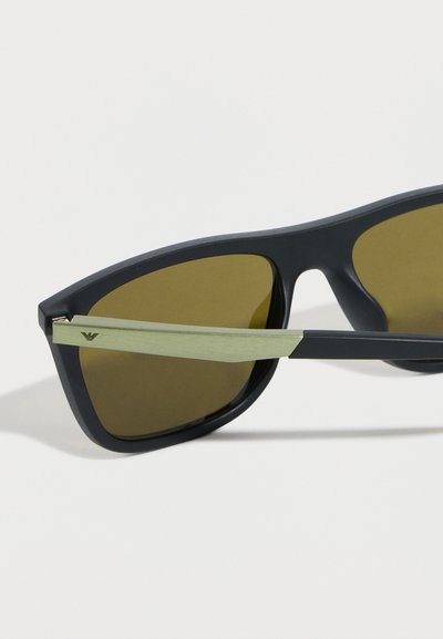 Γυαλιά ηλίου με μαύρο πλαίσιο, φακούς με καφέ απόχρωση και μεταλλικούς βραχίονες σε χρυσή απόχρωση που φέρουν μικρό λογότυπο κοντά στη άρθρωση.