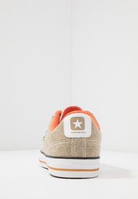 Beżowe płócienne trampki z pomarańczowym wykończeniem i naszywką z logo z białą gwiazdą. Czarne wykończenie i biała gumowa podeszwa z pomarańczowymi i szarymi paskami.
