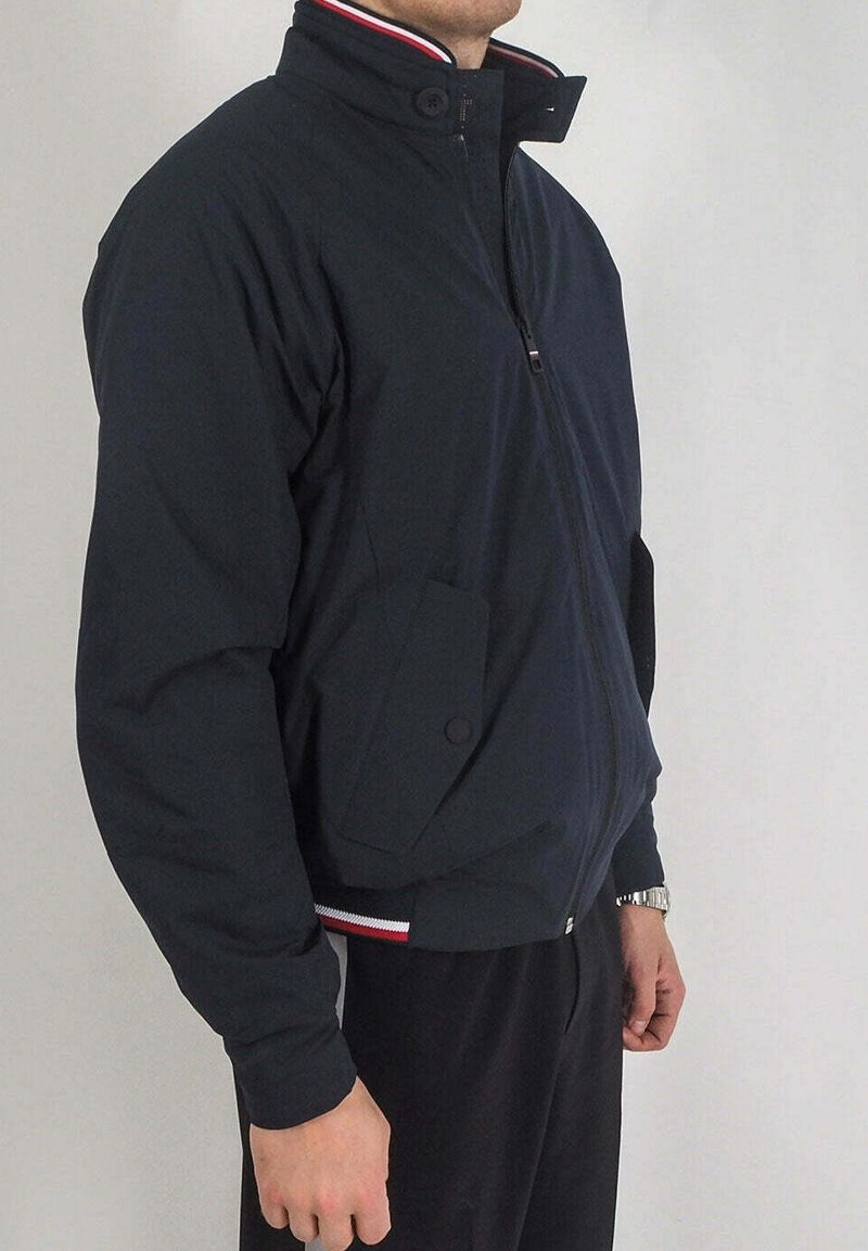 Homme portant une veste bleu marine foncé zippée avec un col montant et des poches latérales, debout devant un fond clair uni.