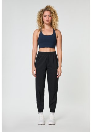 Göndör szőke hajú nő tengerészkék sportmelltartót, fekete melegítőnadrágot és fehér tornacipőt visel, sima világos háttér előtt áll.