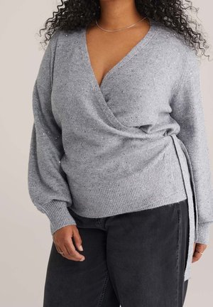 Frau trägt einen glitzernden hellgrauen Wickelpullover mit seitlicher Schleife und schwarze Hose, obere Körperhälfte und entspannte Handhaltung sichtbar.