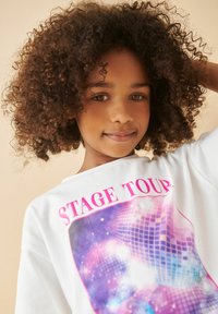 Valge t-särk, millel on värvikas graafiline disain ja tekst "STAGE TOUR" ning diskopalli muster roosade ja lillade toonide kombinatsioonis.