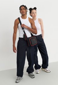 Schwarze Umhängetasche mit roten Akzenten, quadratische Form, abnehmbarer Trageriemen, getragen von einer Person in einem weißen Tanktop und dunkelblauen weiten Jeans.
