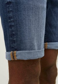 Pantalones cortos de mezclilla en azul oscuro con dobladillo enrollado. La tela presenta una textura sutil, complementada con costuras amarillas contrastantes en el dobladillo.