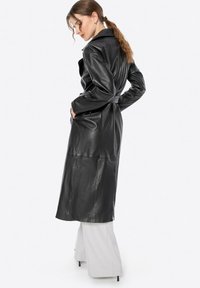 Cappotto trench in pelle nera con vita a cintura, maniche lunghe e tasche laterali. Texture liscia e design sartoriale con un taglio lungo fino al ginocchio.