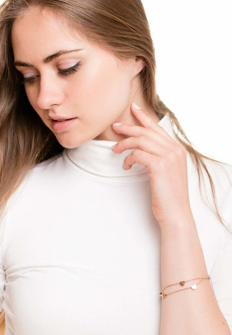 Rose gold bracelet with three small heart charms on a delicate chain, worn on a model's wrist. White turtleneck top. Plain background.