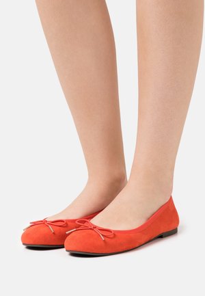 Voeten met felrode suède ballerina's met kleine decoratieve strikjes, afgebeeld tegen een effen witte achtergrond.
