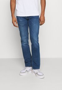 Jeans de mezclilla de color azul oscuro, con un corte recto, un ligero desgaste y un diseño clásico de cinco bolsillos, combinados con zapatillas blancas.