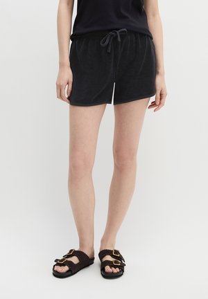 Person iført sorte shorts med snøre, sort top og mørkebrune sandaler med to spænder på hver, stående mod en ensfarvet lys baggrund.