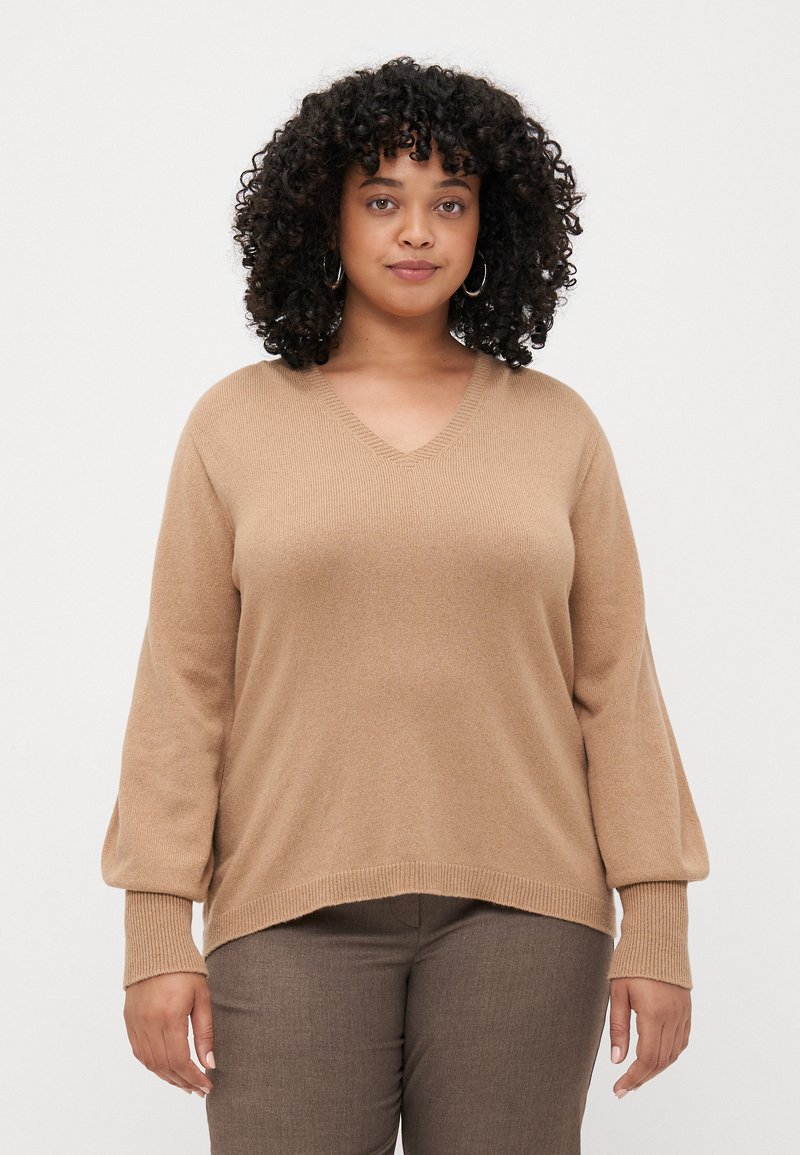 Pull en V beige avec une coupe loose et des poignets ribbinés. Tissu doux avec une texture lisse, présenté associé à un pantalon marron.