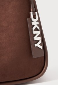 Borsa a spalla in suede marrone con rifiniture in pelle liscia. Presenta un'etichetta con il logo "DKNY" in argento attaccata alla tracolla. Forma arrotondata, design minimale.