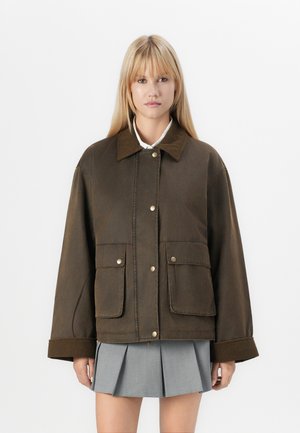 Veste marron surdimensionnée avec un col, dotée de boutons-pression, de deux poches sur le devant et de poignets côtelés. Portée par-dessus une jupe plissée grise.