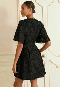 Juoda nėriniuota suknelė su trumpomis, platėjančiomis rankovėmis, prisitaikiusiu liemeniu ir gėlių siuvinėjimu. Atsipalaidavęs siluetas, platėjančia sijonu ir užtrauktuku nugaros dalyje.