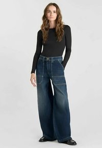 Jean denim à jambes larges de couleur bleu foncé, avec de grandes poches avant, des surpiqûres jaunes, et une taille haute.