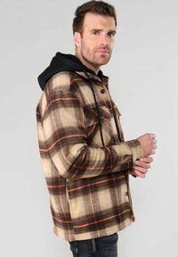 Veste en flanelle à carreaux marron et beige avec une capuche noire. Présente des boutons, des poches latérales et une coupe décontractée. Tissu texturé avec des accents orange.