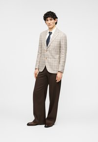 Άνδρας που στέκεται φορώντας μπεζ καρό σακάκι, λευκό πουκάμισο, σκούρα μπλε γραβάτα, φαρδιά καφέ παντελόνια και καφέ δερμάτινα παπούτσια πάνω σε λευκό φόντο.