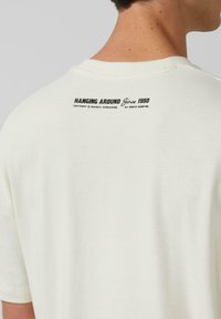 Weißes Baumwoll-T-Shirt mit rundem Halsausschnitt und kurzen Ärmeln. Auf der Rückseite steht in schwarzem Text "HANGING AROUND Since 1950." Glatte Textur.