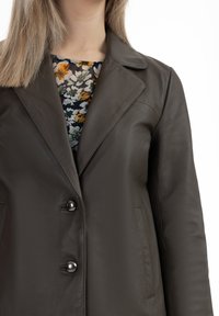 Donkerbruin leren jack met een uitgesneden kraag, twee voorzakken en zwarte knopen. Onder het jack is een blouse met een bloemendessin zichtbaar.