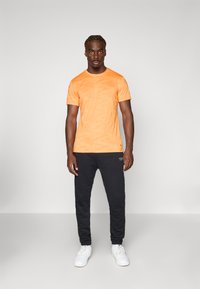 T-shirt laranja de manga curta com um padrão subtil, combinado com calças de treino pretas e ténis brancos, criando um look atlético casual.