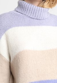 Pull en maille épaisse avec un col haut en côtes. Il présente de larges rayures horizontales de couleur violet clair, blanc et beige. Texturé doux.