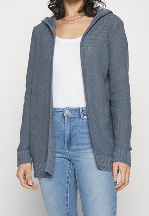 Femme portant un cardigan à capuche gris tricoté par-dessus un débardeur blanc et un jean taille haute bleu clair, debout devant un fond uni.