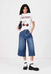 Balts T-krekls ar sarkano "CHERRY" tekstu un divām ķiršu grafikām, pārī ar platām zilu džinsu kulotēm un melnām platformas kurpēm.