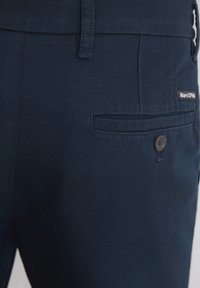 Navyblauwe broek gemaakt van een katoenmix stof. Voorzien van een achterzak met een knoopsluiting en een merklabel. Textuur is zichtbaar.