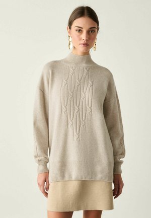 Jeune femme aux cheveux foncés porte un pull beige à torsades avec col montant et manches longues, associé à une jupe beige assortie et des boucles d’oreilles dorées.