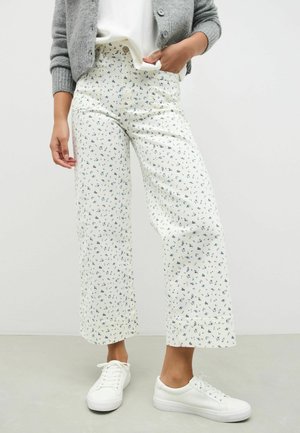 Vrouw draagt witte wijde broek met een kleine blauwe bloemenprint, witte sneakers, wit topje en grijze cardigan, staand tegen een effen achtergrond.