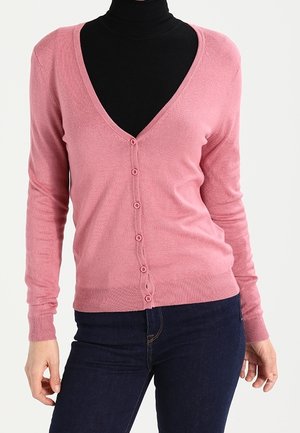 Kvinde iført en tætsiddende lyserød knappet cardigan over en sort højhalset trøje, kombineret med mørkeblå jeans.