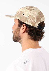 Casquette beige avec un design à motifs comportant des cercles et des triangles. Matière en tissu, visière courbée, sangle réglable à l'arrière.