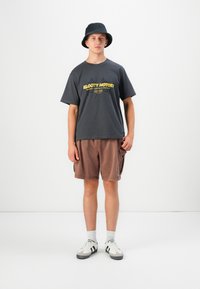 En ung mand iført en sort bucket hat, en mørkegrå t-shirt med grafik, brune cargoshorts, hvide sokker og sorte og hvide sneakers, der står foran en hvid væg.