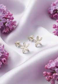 Orecchini a bottone a forma di fiore d'argento posizionati su raso lavanda, circondati da fiori di lilla color viola pallido. Design floreale strutturato.