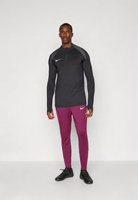 Zwart shirt met lange mouwen en een halve rits, met grijze accenten, gecombineerd met strakke bordeauxrode broek. Beide hebben Nike-logo's in contrasterende kleuren.