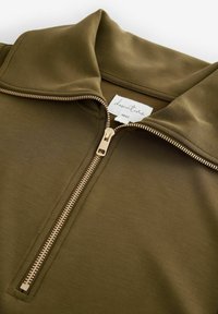 Olijfgroene rits-topt gemaakt van een gladde stof, met een staande kraag en een gouden rits met een rechthoekige trekker. Merkenlabel zichtbaar.