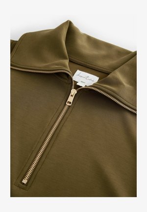 Top cu fermoar de culoare verde măsliniu, realizat dintr-un material neted, având un guler înalt și un fermoar de culoare aurie cu un tab de prindere rectangular. Eticheta brandului este vizibilă.