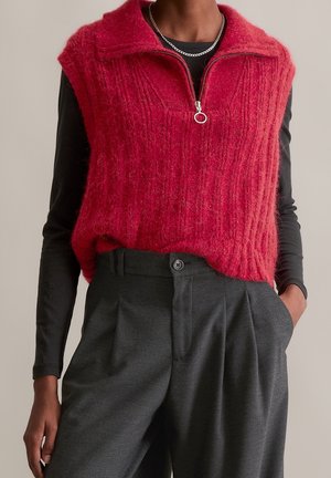 Personne portant un gilet sans manches rouge en tissu duveteux avec fermeture éclair par-dessus une chemise noire à manches longues et un pantalon gris foncé taille haute.