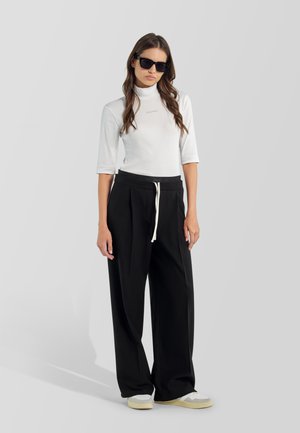 Mujer con gafas de sol negras, top blanco de cuello alto, pantalones negros sueltos con cordón blanco y zapatillas blancas, de pie frente a un fondo liso.