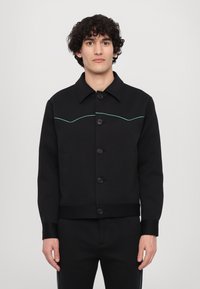 Černá bunda s límcem, předními kapsami a knoflíky. Obsahuje kontrastní zelené prošívání v zakřiveném designu na hrudi.