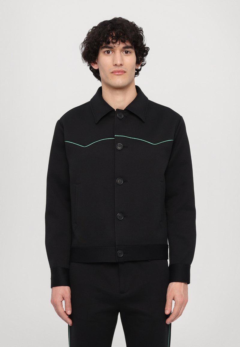 Black jacket with a collar, front pockets, and buttons. Features contrasting green stitching in a curved design along the chest.