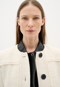 Giacca bianca con texture e colletto in pelle nera, dotata di zip frontale e due bottoni neri sul petto.