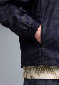 Personne portant une veste sombre à motifs avec un poignet élastique, main dans la poche, et une chemise beige à motifs en dessous, sur fond uni.