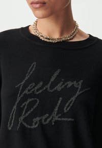 Maglione nero a maglia con stampa argentata metallizzata "Feeling Rock". Collana a catena dorata multi-strato. Vista ravvicinata.